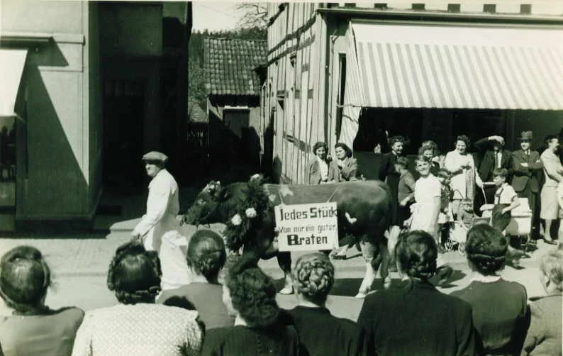 Fleischer Ernst Neise führt einen Bullen vor. Der Bulle ist festlich geschmückt und trägt ein Schild "Jedes Stück von mir ein guter Braten". Im Hintergrund ist zwischen den Häusern "ehem. Grosche" und "Modehaus Humpert" die Schmiede zu sehen.